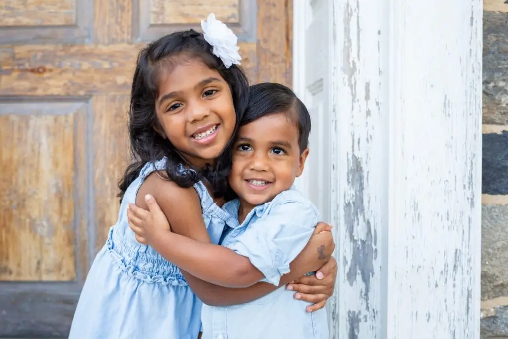 Brother and sister hug affectionately by an old stone home.